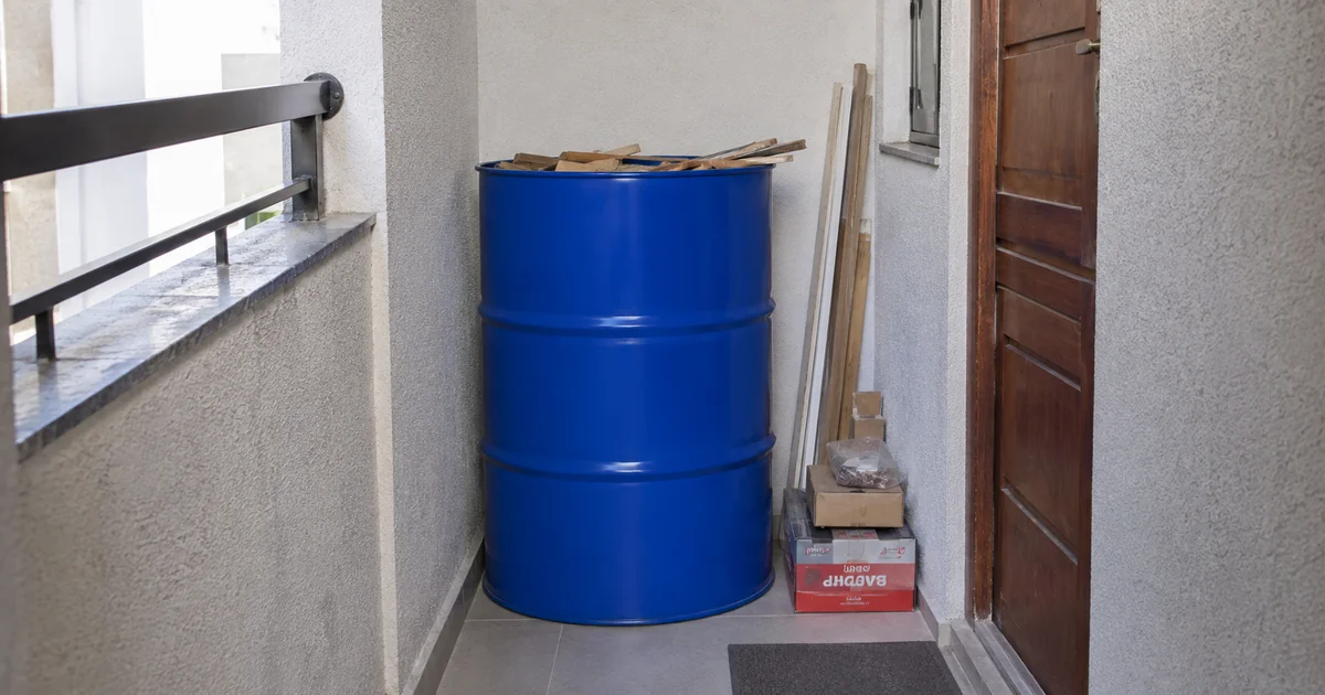 Tambor metálico azul de 200 litros posicionado em corredor estreito de apartamento, próximo à entrada, de forma organizada